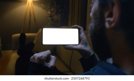 Man lying on couch using smartphone horizontally with chroma key white screen. Male watching series or video at home at night.