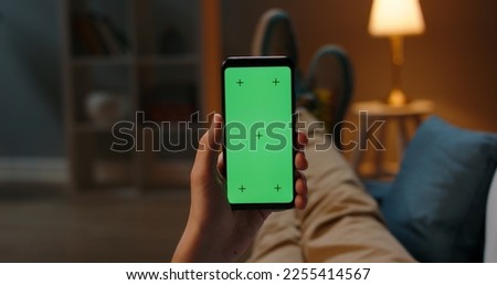 Man lying on couch using smartphone with chroma key green screen at night, scrolling through social media or online shop - internet, communications concept close up 