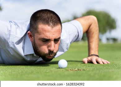Man lying on a golf course blowing a golf ball into a hole
