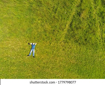 Man lying on the ground in a park on a green grass field. Man dressed in blue t shirt and jeans hands behind hes head. Aerial top down view.