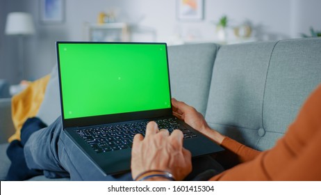 Man Lying on His Sofa At Home, Uses Laptop with Green Mock-up Screen. Over The Shoulder Shot of Anonymous Man in the Living Room. - Powered by Shutterstock