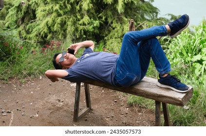 Man lying on old wooden bench over hilltop making a call on his smartphone