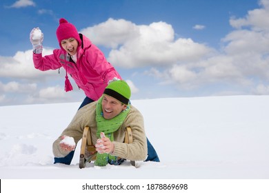 A Man Lying On Sled With His Cute Girlfriend Standing Above Trying To Playfully Hit Him With A Big Snow Ball