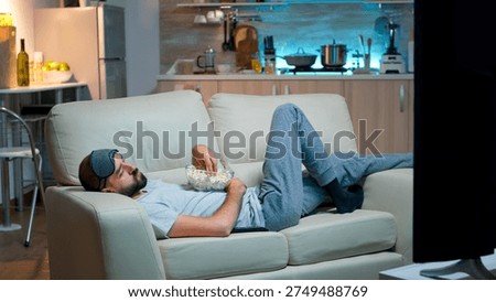 Man lying on sofa, falling asleep in fron of the TV with popcorn bowl in his hands. Tired alone man in living room after a hard day at work