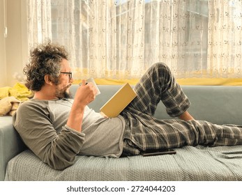 A man lying on the sofa reading a book. My Sunday routine: a book and coffee. A middle-aged man with curly hair and glasses is resting. Model, people, human. Horizontal photo.