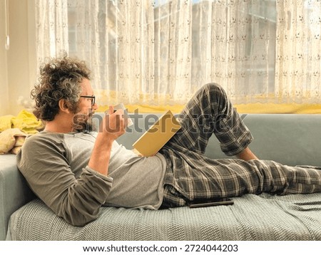 A man lying on the sofa reading a book. My Sunday routine: a book and coffee. A middle-aged man with curly hair and glasses is resting. Model, people, human. Horizontal photo.