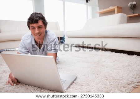 Man lying on stomach using silver laptop on shag rug in bright living room, wearing polo. Person, home, remotework, notebook, minimalist, modern, neutral