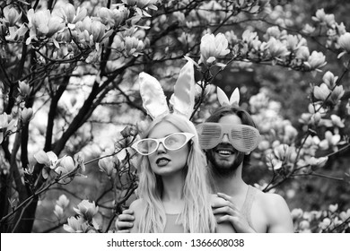 Man Or Macho Hugging Angry Girl Or Pretty Woman On Floral Environment. Funny Couple, Girlfriend And Boyfriend In Glasses And Rosy, Bunny Ears In Blossoming Magnolia Garden. Spring. Easter