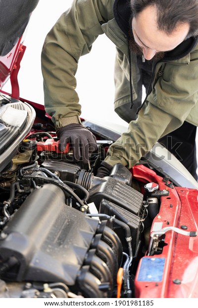 Man makes car repairs, change battery.\
Worker repairing car under red hood changes\
parts