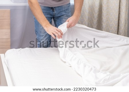 A man makes his cozy bed with fresh bright white bed linen. Making the bed with fresh bed linen by a white man. The day of the change of bedclothes. The day of washing bedclothes in the laundry room.