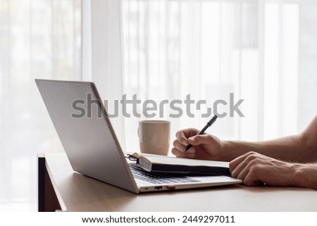 a man makes notes in a notebook , in front of a laptop at a small table, image in light colors, online lesson or online meeting, cafe or office, modern silver laptop