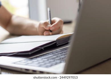  a man makes notes in a notebook ,typing in laptop ,at a table, image in light colors, online lesson or online meeting, cafe or office, modern silver laptop, blurred window background