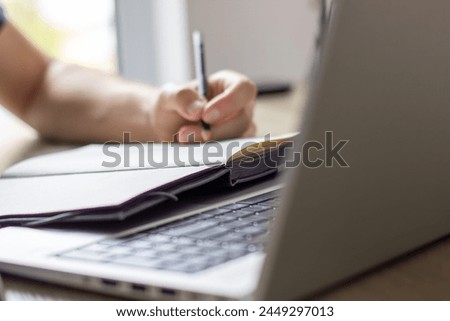  a man makes notes in a notebook ,typing in laptop ,at a table, image in light colors, online lesson or online meeting, cafe or office, modern silver laptop, blurred window background