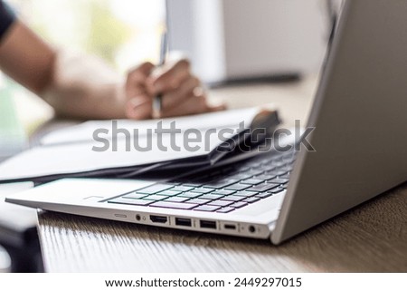  a man makes notes in a notebook ,typing in laptop ,at a table, image in light colors, online lesson or online meeting, cafe or office, modern silver laptop, blurred window background