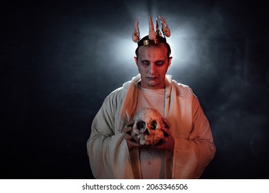 a man in the makeup of a dead man puts a crown with skulls on his head. a gloomy gothic image for Halloween. creative photography in the studio with smoke and light. a demon in a white robe