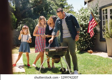 Man making barbeque in the backyard standing with his family members. Family celebrating an occasion together with the american flag in the background.