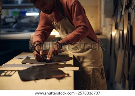 Man making leather items in a workshop, examining details carefully, wearing protective gear and apron while using different tools during a design process