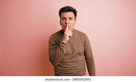 Man making a shushing gesture, requesting silence or discretion with his finger on his lips on a pink background