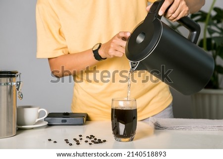 Man making use kettle pouring hot water making iced coffee or cold americano coffee into the cup with equipment, tool digital scale on the wooden bar at kitchen home