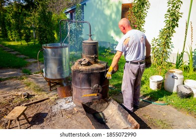 Man Is Manually Turning Lever To Mix Fruit Marc In Boiler Of Homemade Distillery Made Of Copper, Making Moonshine Schnapps, Alcoholic Beverages Such As Brandy, Cognac, Whiskey, Bourbon, Gin, Scotch.