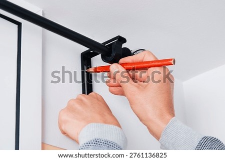 a man marks the drilling point on a white wall with a red pencil while holding a black metal bracket for a curtain rod in place near the corner of the room