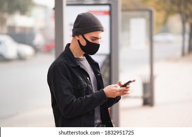 Man In Mask With A Cell Phone At A Bus Stop. Millennial Using Smartphone At Public Place, Covid Time