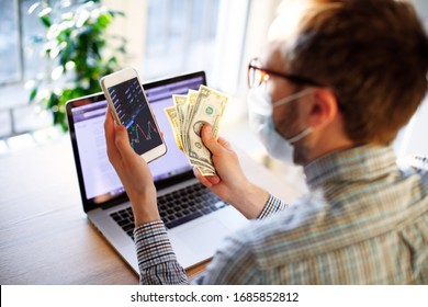 Man in mask at coronavirus quarantine isolation working remotely at home, counting money savings due to economic financial crisis and unemployment looking at phone with currency rates, income decrease