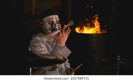 Man in mask plays rock drums. Clip. Masked drummer is actively playing on background of fire in forest. Masked man plays drums at festival in night forest - Powered by Shutterstock