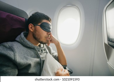 Man In A Mask For Sleeping, Sits In The Airplane's Chair Near The Porthole. Rest And Comfortable Flight In The Plane.