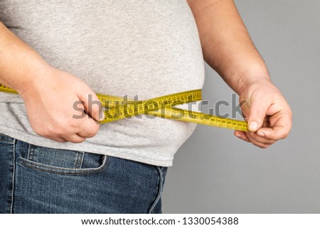 A man measures his fat belly with a measuring tape. on a gray background