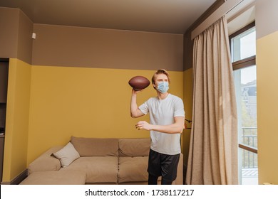 Man In Medical Mask Holding American Football In Living Room