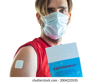 A man in a medical mask holds a vaccination certificate in his hand. The shoulder where the injection was sealed with a plaster. Translation: certificate of vaccination against COVID-19