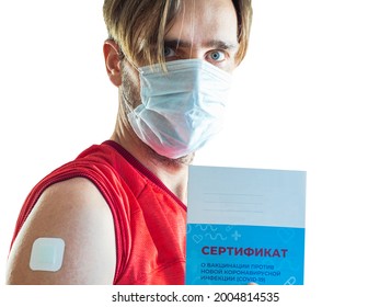 A man in a medical mask holds a vaccination certificate in his hand. The injection site on the shoulder is sealed with a plaster. Translation: certificate of vaccination against COVID-19