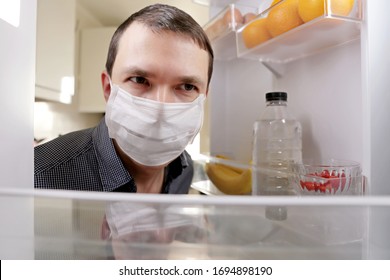 Man In A Medical Mask Looks Into The Fridge With Food. Concept Of Diet, Obesity During Home Quarantine At Covid-19 Coronavirus Pandemic