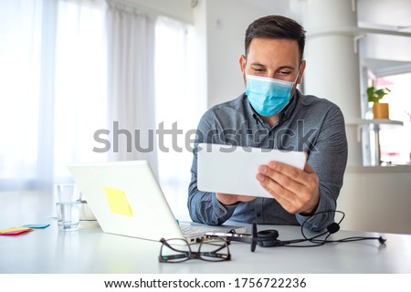 Man in a medical mask at the office. Man works remotely. The guy uses a tablet PC and a laptop for work, and medicine mask and antiseptic for self protective. Coronavirus pandemic, influenza, covid19