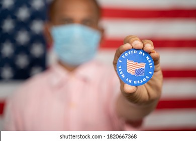 Man In Medical Mask Showing I Voted By Mail Sticker With US Flag As Background - Concept Of Mail In Voting At USA Election