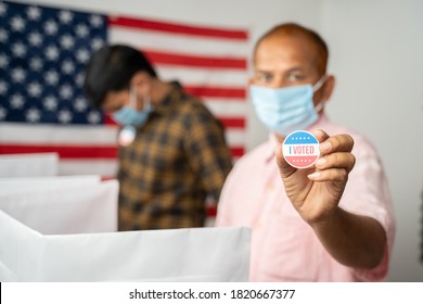 Man In Medical Mask Showing I Voted Sticker At Polling Booth With US Flag As Background - Concept In Person Voting At US Election