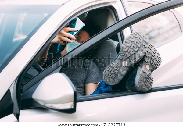 A man in a medical mask sits in a car with his\
legs upstairs. Man in protective mask takes a selfie on a cell\
phone in the car. Protection from coronavirus epidemic pandemic,\
covid-19. New reality