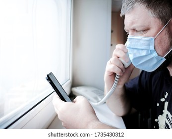 A Man In A Medical Mask Sits In Front Of A Window And Talks On A Landline Phone, Holding A Smartphone In His Hand. He Worries About Family Health During The Quarantine Of The Coronavirus Epidemic.