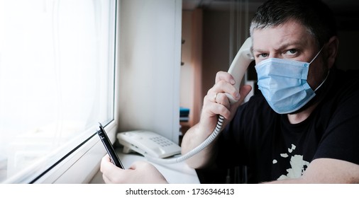 A Man In A Medical Mask Sits In Front Of A Window And Talks On A Landline Phone, Holding A Smartphone In His Hand. He Worries About Family Health During The Quarantine Of The Coronavirus Epidemic.