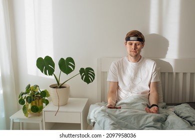 Man Meditating On Bed.  Young Male Sitting And Relax In White Bedroom. Guided Morning Meditation. Meditation App And Wearable Technology. Headband For Sleeping And For Health Tracking. Calm Lifestyle