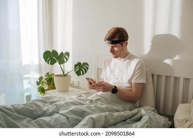 Man Meditating On Bed. Young Male Sitting And Relax In White Bedroom. Audio Guided Morning Meditation App. Male Listening Calm Audio In Headphones And Wearable Technology. Headband For Sleeping. 