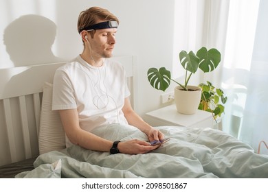 Man Meditating On Bed. Young Male Sitting And Relax In White Bedroom. Audio Guided Morning Meditation App. Male Listening Calm Audio In Headphones And Wearable Technology. Headband For Sleeping. 