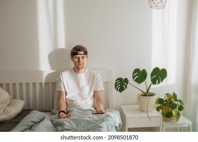 Man Meditating On Bed. Young Male Sitting And Relax In White Bedroom. Audio Guided Morning Meditation App. Male Listening Calm Audio In Headphones And Wearable Technology. Headband For Sleeping. 