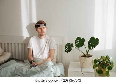 Man Meditating On Bed. Young Male Sitting And Relax In White Bedroom. Guided Morning Meditation. Meditation App And Wearable Technology. Headband For Sleeping And For Health Tracking. Calm Lifestyle