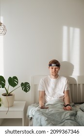 Man Meditating On Bed. Young Male Sitting And Relax In White Bedroom. Guided Morning Meditation. Meditation App And Wearable Technology. Headband For Sleeping And For Health Tracking. Calm Lifestyle