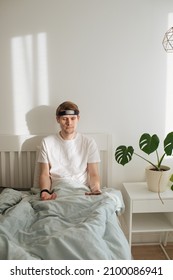 Man Meditating On Bed. Young Male Sitting And Relax In White Bedroom. Guided Morning Meditation. Meditation App And Wearable Technology. Headband For Sleeping And For Health Tracking. Calm Lifestyle