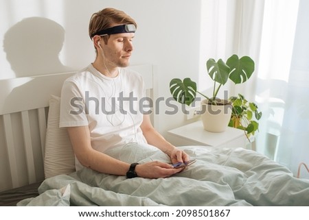 Man meditating on bed. Young male sitting and relax in white bedroom. Audio guided morning meditation app. Male listening calm audio in headphones and wearable technology. Headband for sleeping. 