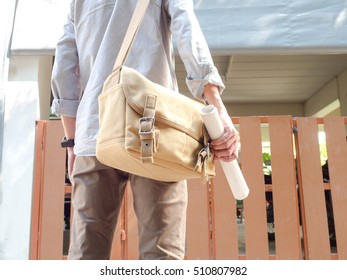 A Man With Messenger Bag Standing In Front Of The House And Holding Roll Paper