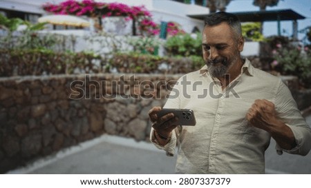 Man middle aged hispanic with grey hair and beard holding smartphone and pumping fist on street near stone wall building; joy celebration victory.